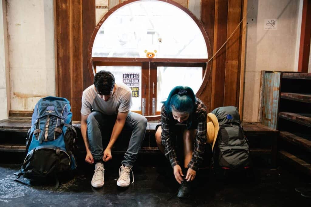 A man and woman sit indoors tying shoes next to backpacks, ready for travel.